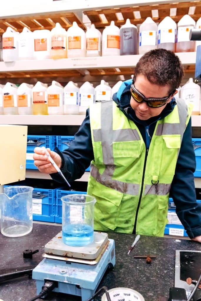 Dr Adam Hunniford founder of PiP Chemicals in Newtownards standing in the warehouse beside a shelf full of chemicals containers, Dr Hunniford is wearing a high vis vest