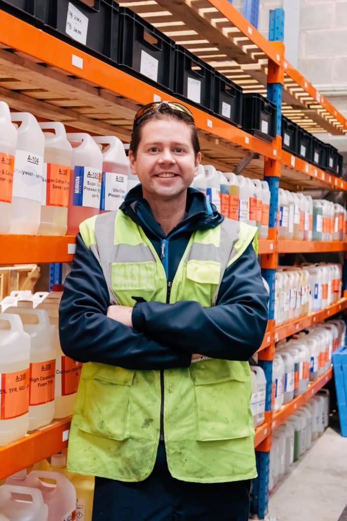 Dr Adam Hunniford founder of PiP Chemicals in Newtownards standing in the warehouse beside a shelf full of chemicals containers, Dr Hunniford is wearing a high vis vest