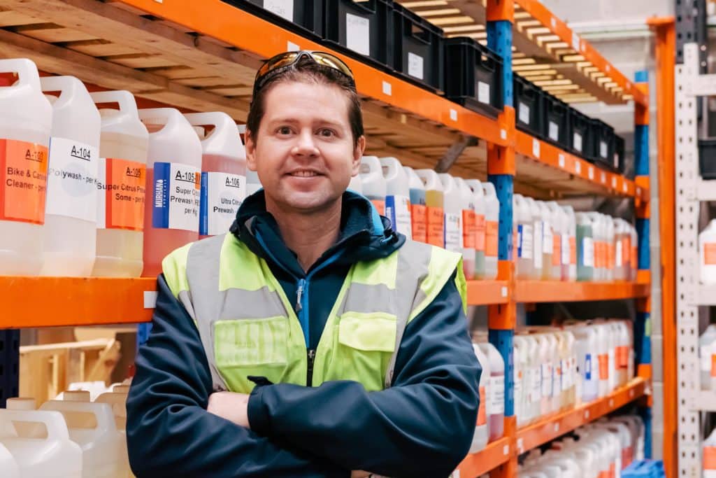 Dr Adam Hunniford founder of PiP Chemicals in Newtownards standing in the warehouse beside a shelf full of chemicals containers, Dr Hunniford is wearing a high vis vest