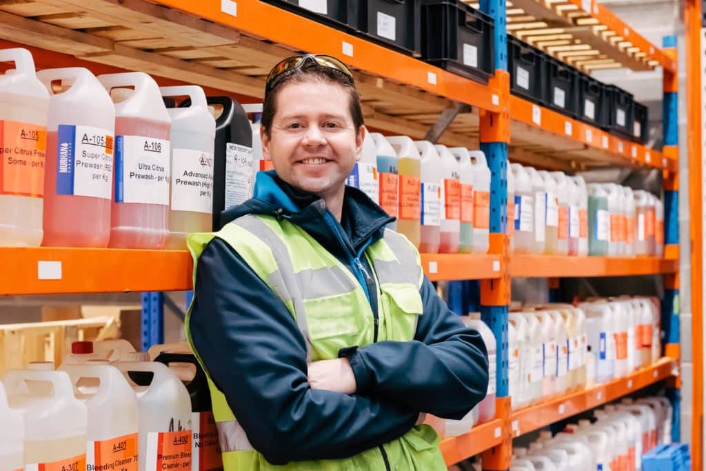 Dr Adam Hunniford founder of PiP Chemicals in Newtownards standing in the warehouse beside a shelf full of chemicals containers, Dr Hunniford is wearing a high vis vest